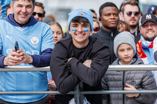 A group of Manchester City fans celebrating, one fan with face paint and Manchester City hat is prominently smiling and leaning on a railing.