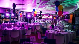 A festive ballroom event with round tables set for dining, colorful balloons, and vibrant lighting creating a celebratory ambiance.
