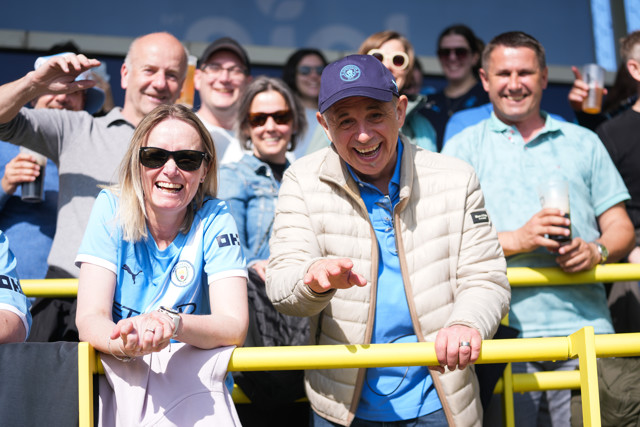 A group of Manchester City fans are excited and smiling at a football match, wearing team jerseys and holding drinks.