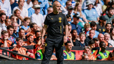 A man in a black sports tracksuit stands on the sidelines of a football match observing the field. The crowd in the background is filled with spectators wearing light blue and white clothing.