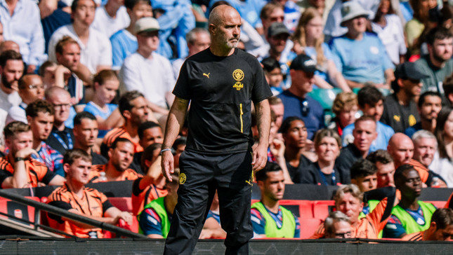 A man in a black sports tracksuit stands on the sidelines of a football match observing the field. The crowd in the background is filled with spectators wearing light blue and white clothing.