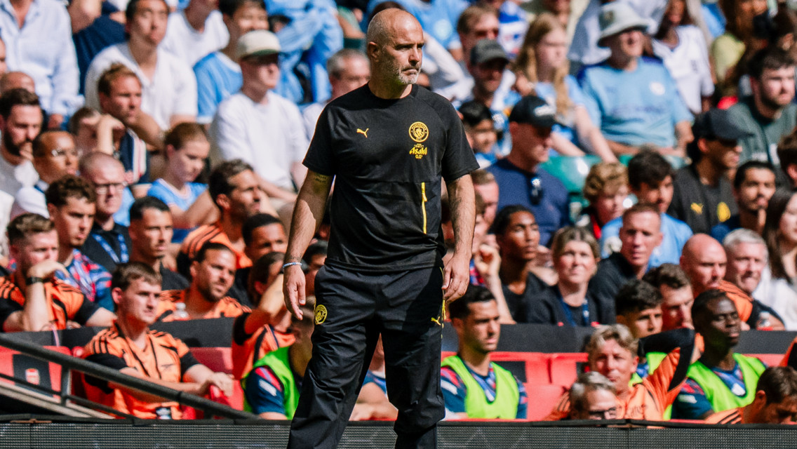 A man in a black sports tracksuit stands on the sidelines of a football match observing the field. The crowd in the background is filled with spectators wearing light blue and white clothing.