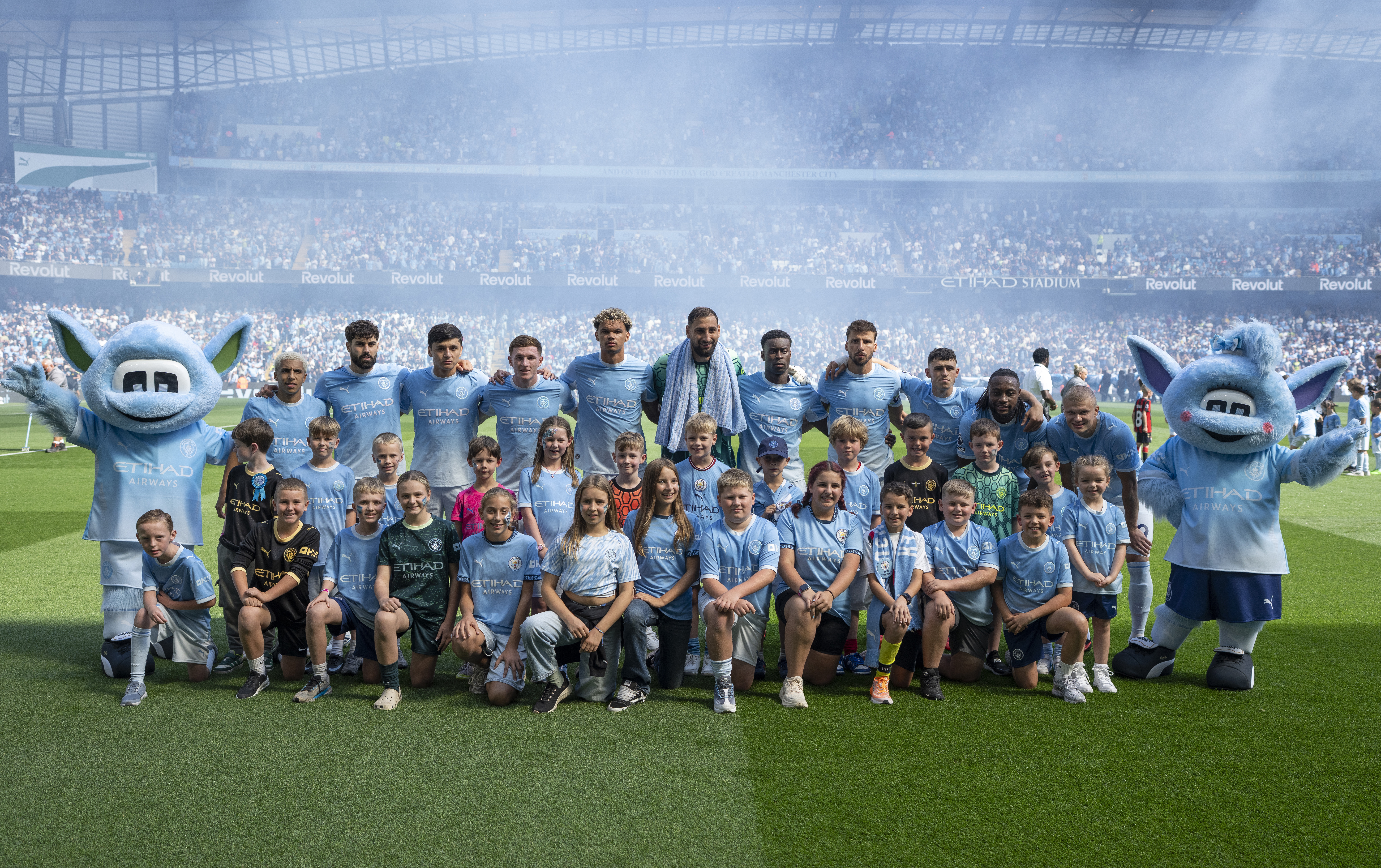 A group photo of Manchester City players with young player escorts and two team mascots at Etihad Stadium.