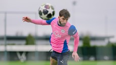 A soccer player wearing a pink and blue Manchester City jersey heads a soccer ball during practice on a field.