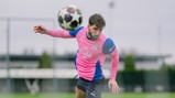 A soccer player wearing a pink and blue Manchester City jersey heads a soccer ball during practice on a field.