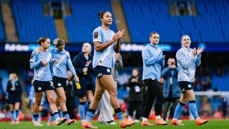 Women’s football team in light blue jerseys clapping as they walk on a football field. Empty blue stadium seats are visible in the background.