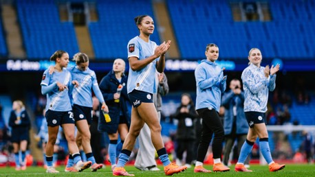 Women’s football team in light blue jerseys clapping as they walk on a football field. Empty blue stadium seats are visible in the background.