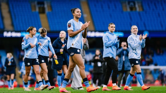 Women’s football team in light blue jerseys clapping as they walk on a football field. Empty blue stadium seats are visible in the background.