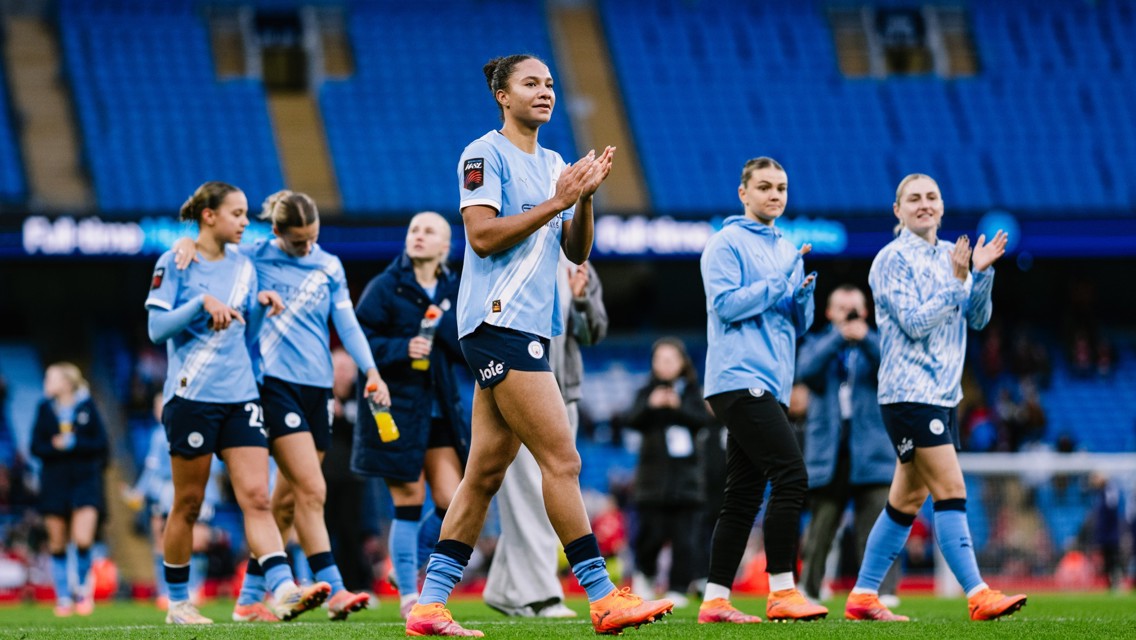 Women’s football team in light blue jerseys clapping as they walk on a football field. Empty blue stadium seats are visible in the background.