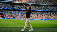 Person with raised arms on a football field with a packed stadium in the background.
