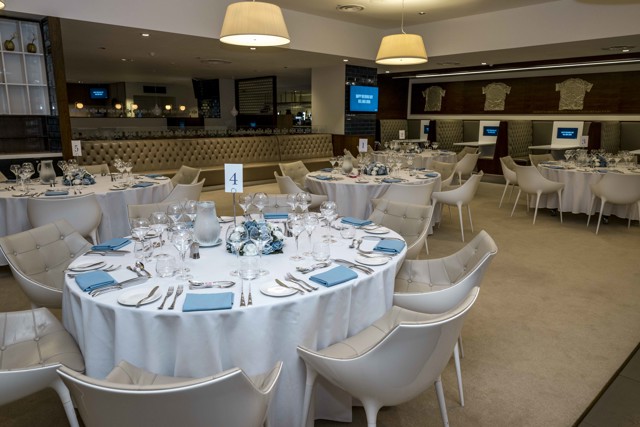 A banquet hall set up for an event with round tables covered in white tablecloths, each adorned with blue napkins, glassware, and floral centerpieces. There are numbered table signs and modern, white chairs arranged around the tables.