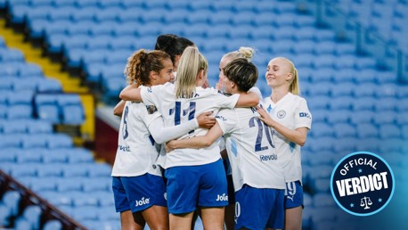 A group of football players in white jerseys and blue shorts celebrating a goal on a blue seated stadium background, with an 'Official Verdict' badge in the corner.