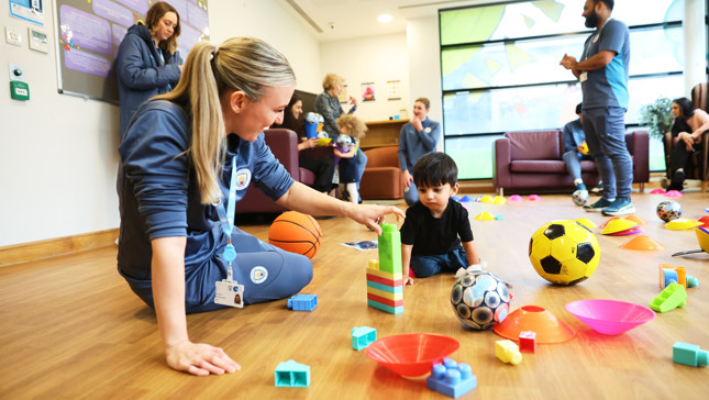 A caregiver and a child are playing with building blocks surrounded by various toys on a wooden floor in a community center.