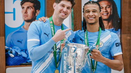 Two Manchester City players in a light blue kit celebrate with the Carabao Cup trophy and medals.