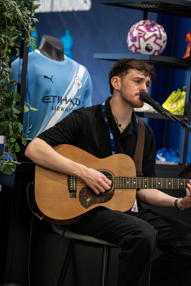 A person with a guitar performing at an event. A Manchester City jersey and football are visible in the background.