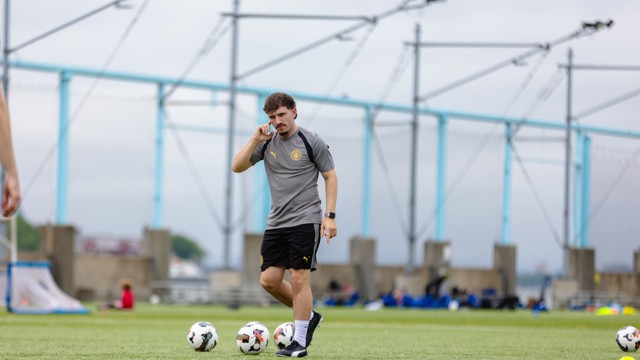 A person in a grey sports shirt and black shorts is on a soccer field with multiple soccer balls and goalposts in the background.