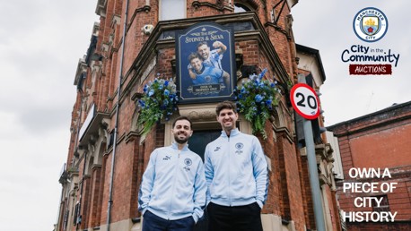 Two people in Manchester City attire stand in front of a pub named 'The Stones & Silva' with a Manchester City logo visible.