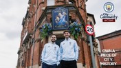 Two people in Manchester City attire stand in front of a pub named 'The Stones & Silva' with a Manchester City logo visible.