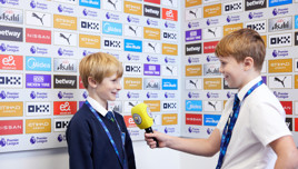 Two young individuals holding press badges engage in a mock interview setup, with one holding a yellow microphone. The background has sponsor logos typical of a football press area, including Premier League logos.