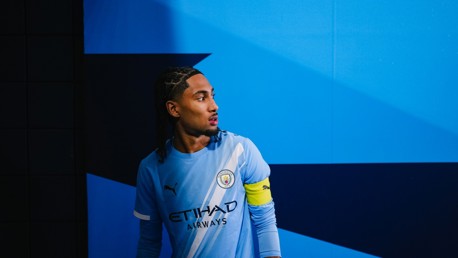A person wearing a Manchester City football kit stands in a blue-themed tunnel with a tile pattern and gradient background.