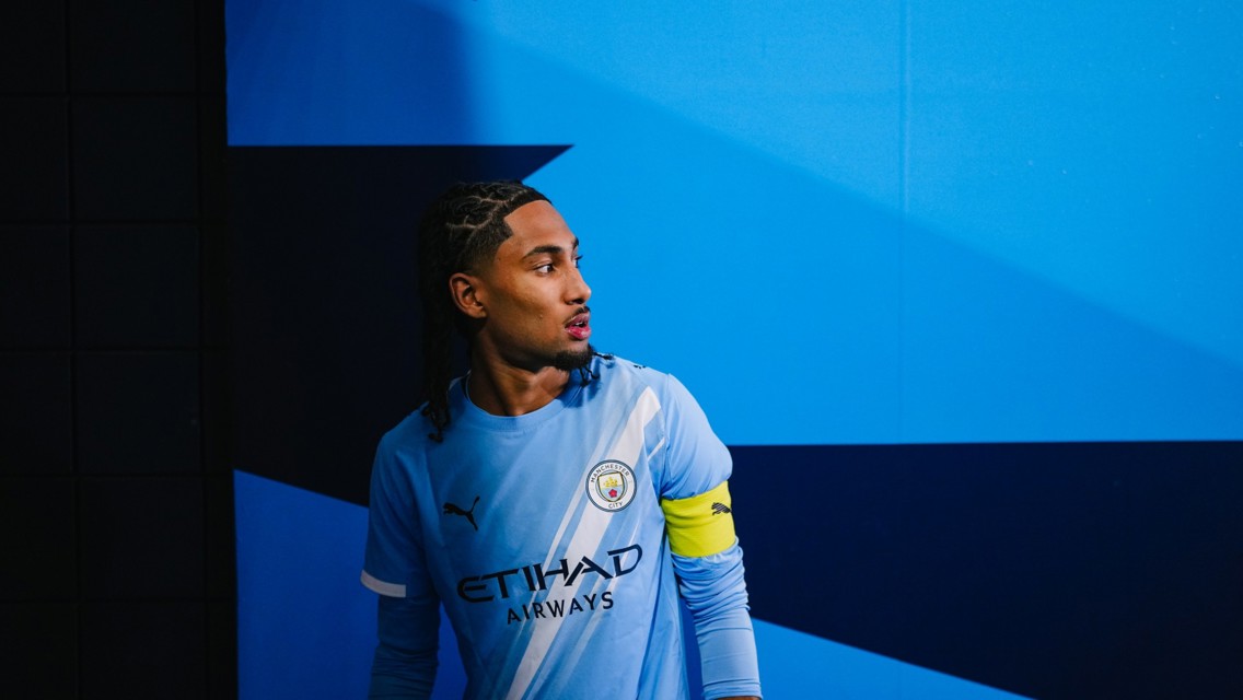 A person wearing a Manchester City football kit stands in a blue-themed tunnel with a tile pattern and gradient background.