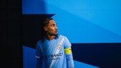 A person wearing a Manchester City football kit stands in a blue-themed tunnel with a tile pattern and gradient background.