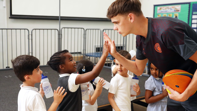 A Manchester City player, wearing the club's logo, engages with schoolchildren in a classroom setting, high-fiving them while holding football training disks. Children hold water bottles and wear matching uniforms.