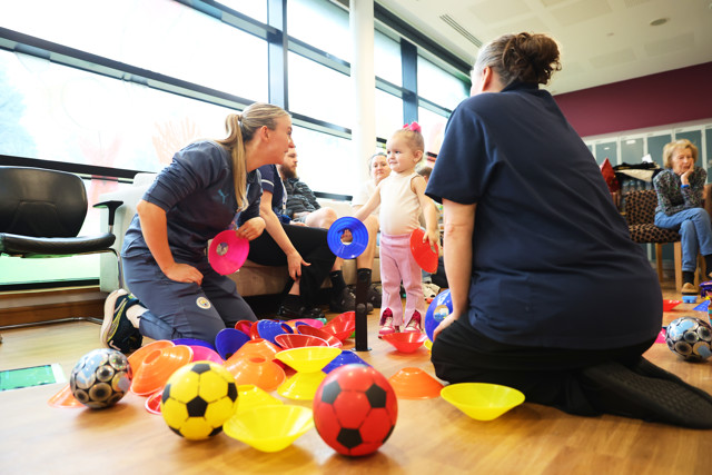A group of women and children playing with colorful cones and footballs on the floor using a variety of colored training cones. There are footballs and training equipment scattered around.