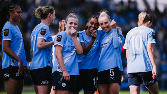 Manchester City women's football players celebrating on the field, wearing the team's light blue kits with sponsor logos.