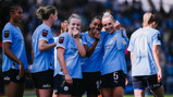 Manchester City women's football players celebrating on the field, wearing the team's light blue kits with sponsor logos.