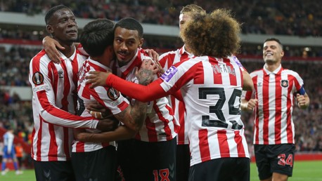 Soccer players in red and white striped uniforms celebrate a goal, huddled together on the field.