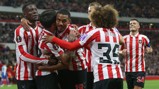 Soccer players in red and white striped uniforms celebrate a goal, huddled together on the field.