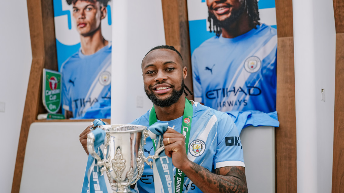Football player in Manchester City kit holding Carabao Cup trophy with team banners in the background.