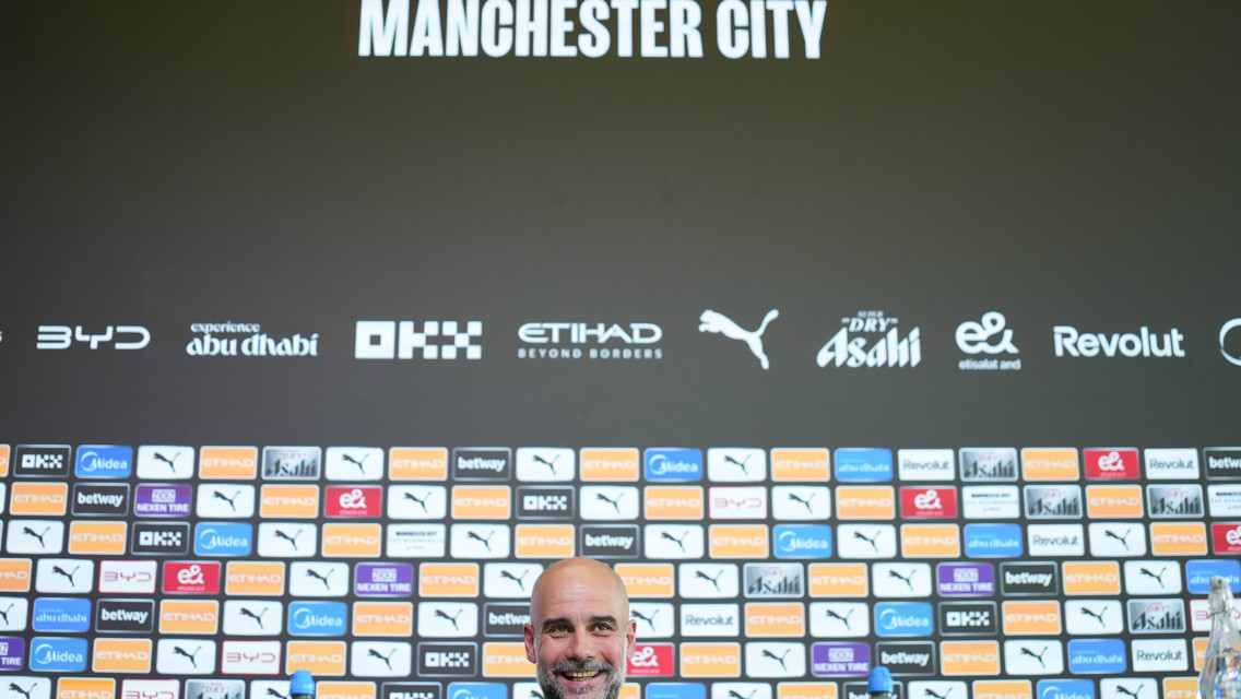 A press conference setting with a Manchester City backdrop featuring various sponsor logos and a blurred person at the podium.