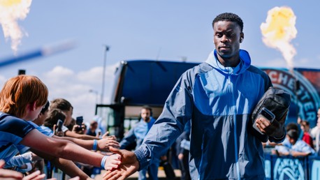 A football player in a Manchester City tracksuit greets fans while holding a phone. Flames are visible in the background near a Man City logo.