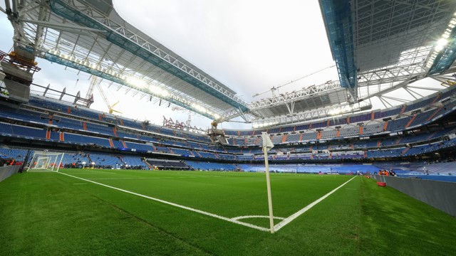 An empty football stadium with green grass, seats, and a partially constructed roof.