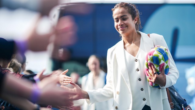 A woman in a white blazer holds a colorful bag while greeting fans and shaking hands outside a bus.