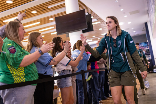 A person in shorts and a jacket high-fives fans behind a barrier, with sports jerseys visible. The environment suggests a sports venue or event.