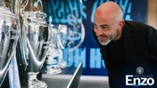 A man in a black jacket is observing a display of football trophies, including the Premier League trophy. The words 'Welcome back Enzo' and Manchester City logo are visible in the lower right corner.