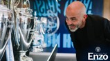 A man in a black jacket is observing a display of football trophies, including the Premier League trophy. The words 'Welcome back Enzo' and Manchester City logo are visible in the lower right corner.