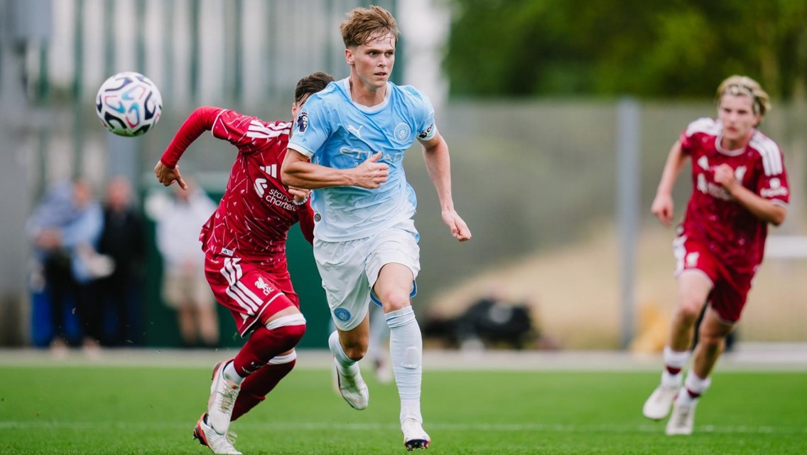 Two soccer players from teams in light blue and red kits compete for the ball on a field during a match.