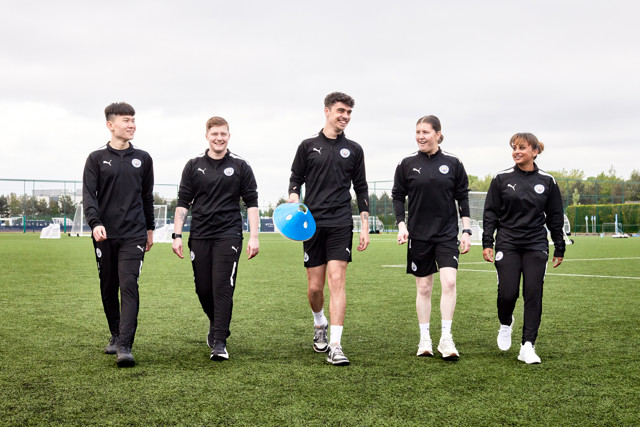 Five Manchester City staff or players wearing training gear walking on a football pitch. One person is holding training equipment.
