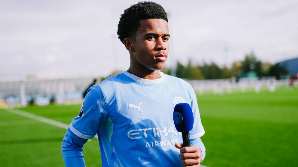 A young footballer wearing a Manchester City jersey holds a microphone on a football pitch.