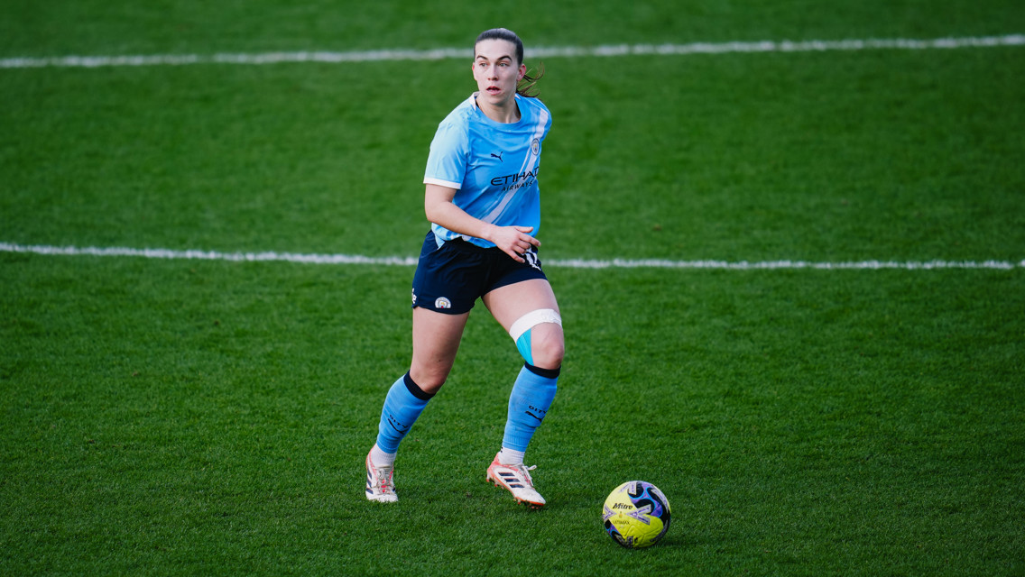 A female soccer player wearing a Manchester City uniform on a field. The player has a knee brace and is in motion near a soccer ball.
