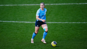 A female soccer player wearing a Manchester City uniform on a field. The player has a knee brace and is in motion near a soccer ball.