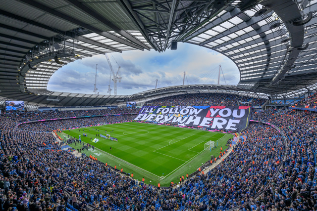 A full view of Etihad Stadium during a football match, with fans displaying a large banner reading 'I'll follow you everywhere'.