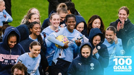 Manchester City Women's team celebrating Khadija 'Bunny' Shaw's achievement of scoring 100 goals, as indicated on the image text. Shaw is holding a ball and a trophy among teammates on the field.