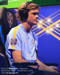 A player wearing headphones and a Manchester City jersey sits in an esports competition setup with 'FIFA eWorld Cup Grand Final 18' branding.