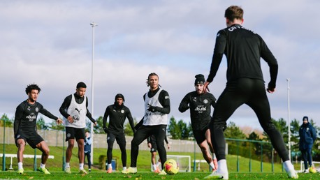 Six Manchester City players in training session, wearing training kits with club logo, practicing on a grassy field with cloudy sky in the background.