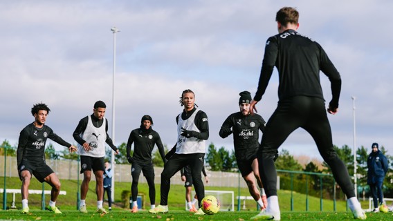 Six Manchester City players in training session, wearing training kits with club logo, practicing on a grassy field with cloudy sky in the background.